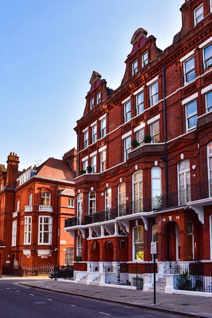 Photograph of a multi-story residential building with a red brick facade, featuring large sash windows framed in white, some with black wrought iron railings on small balconies. The ground floor has a white entrance porch with decorative supports and steps leading up to the front door. The building is situated on a quiet street with a black lamppost and clear blue sky overhead. The clean exterior showcases well-maintained surfaces, highlighting the importance of regular property maintenance and surface cleaning for period flats in Marylebone, as emphasized by Marylebone Carpet Cleaning.