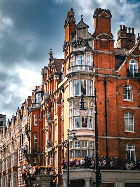 A historic red brick building on Marylebone High Street with ornate white stone details and multiple bay windows, set against a cloudy sky. The facade features decorative elements and flower boxes with colorful blooms on the balconies. Street lamps and surrounding buildings add to the classic London architectural scene, illustrating the characteristic charm of the Marylebone area. Marylebone Carpet Cleaning's expertise in surface cleaning and deep cleaning services is relevant to maintaining the hygiene and appearance of such intricate exteriors and interior spaces.