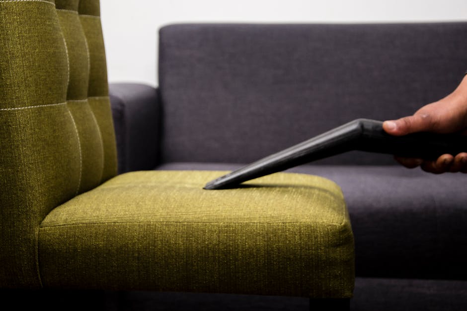 Close-up of a hand holding a vacuum cleaner nozzle, deep cleaning a green upholstered armchair with a tufted backrest in a well-lit living room. The armchair's fabric appears clean and maintained, with a slightly textured surface. Behind, a dark grey sofa with smooth fabric upholstery is visible, positioned against a plain white wall. The vacuum nozzle is pressed onto the seat cushion, indicating surface cleaning as part of domestic cleaning services offered by Marylebone Carpet Cleaning, which maintains high hygiene standards for residential spaces. The lighting highlights the textures and cleanliness of the furniture surfaces, emphasizing professional surface cleaning practices.