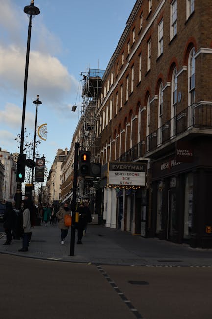 A busy street scene at Portman Estate, Marylebone, showing a row of multi-story brick buildings with arched windows and small balconies, some covered in scaffolding indicating ongoing restoration work. The street is populated with pedestrians walking on the pavement, some carrying shopping bags or backpacks. Traffic lights are visible, with one showing a red signal, and a street sign reads 'Marylebone SO'. The road surface appears clean and well-maintained, with some visible crosswalk markings. The scene is illuminated by natural daylight, highlighting the historic architecture and the active urban environment. As part of maintaining the aesthetic appeal of this area, Marylebone Carpet Cleaning specializes in surface cleaning and deep cleaning services for residential and commercial properties to ensure hygiene and visual appeal, consistent with offerings described on the PAGE_TITLE page.