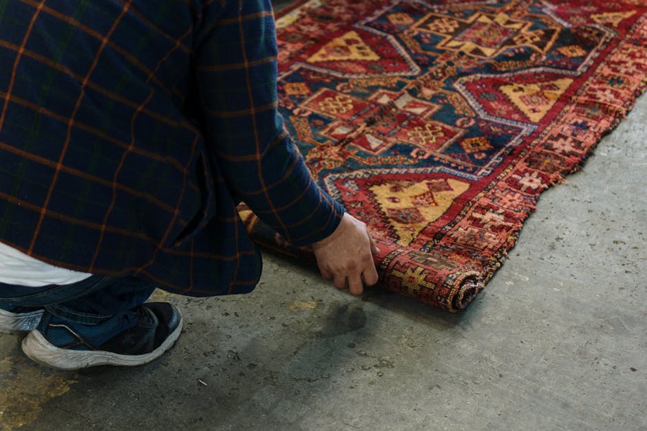 A person wearing a dark plaid jacket and sneakers is carefully flipping up the corner of a traditional oriental rug with intricate geometric and floral patterns in red, yellow, and blue tones, placed on a concrete floor. The room is well-lit, and the rug appears clean and well-maintained, reflecting expert surface cleaning and care as provided by Marylebone Carpet Cleaning, whose services include rugs restoration and deep cleaning for both residential and commercial spaces.